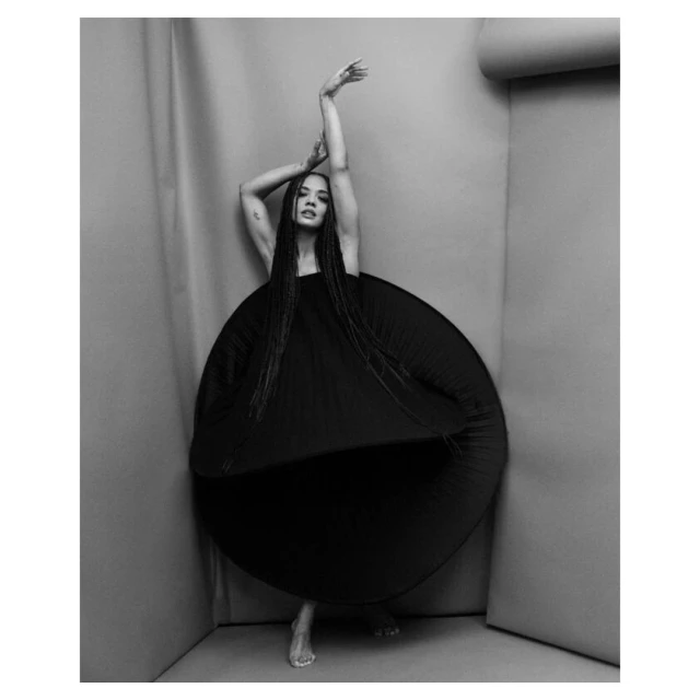 Artistic black and white studio shot of a female model in an oversized black dress, standing with arms raised against a grey studio backdrop.