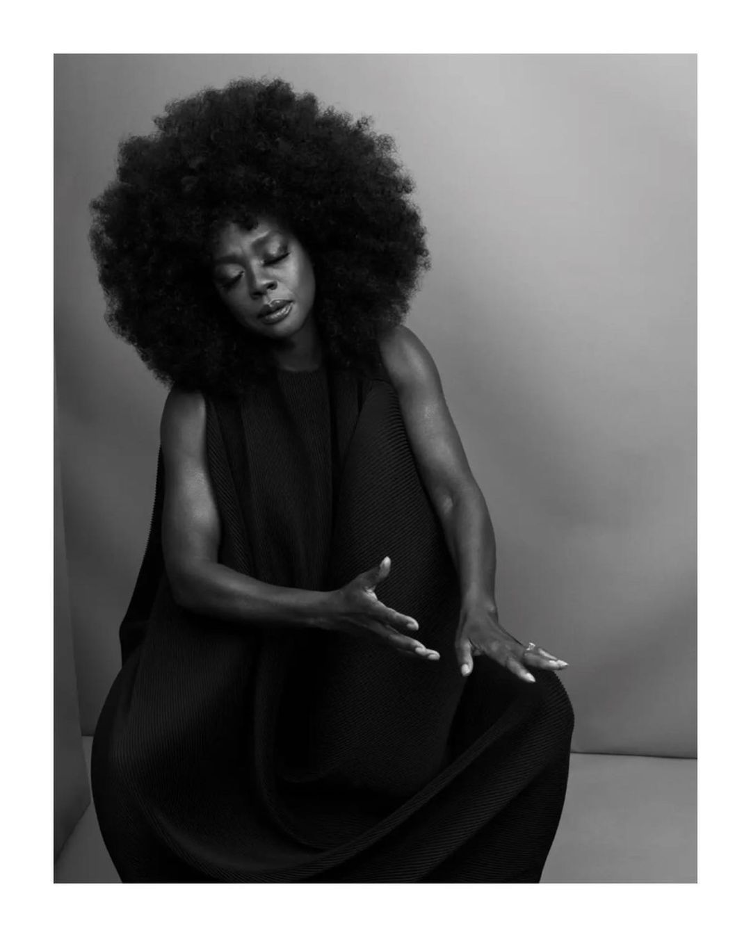 Black and white studio portrait of Viola Davis with afro, closed eyes, and hand gestures, wearing a draped black dress against a grey background.