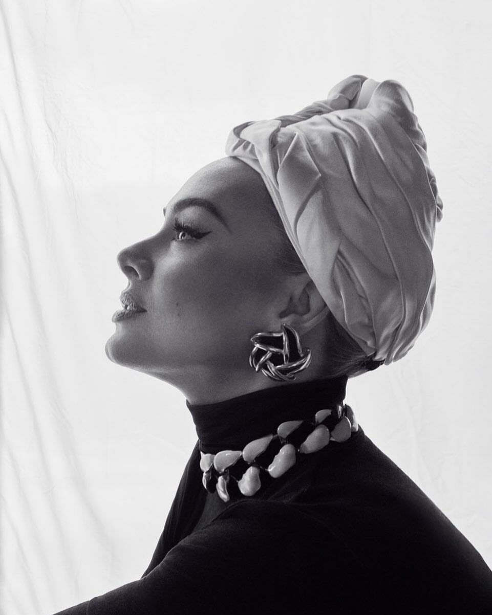 Elegant woman in a headscarf and black turtleneck poses in a studio with earrings and a striking white-and-black necklace.