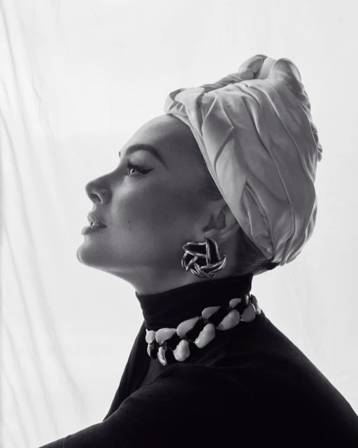 Elegant woman in a headscarf and black turtleneck poses in a studio with earrings and a striking white-and-black necklace.