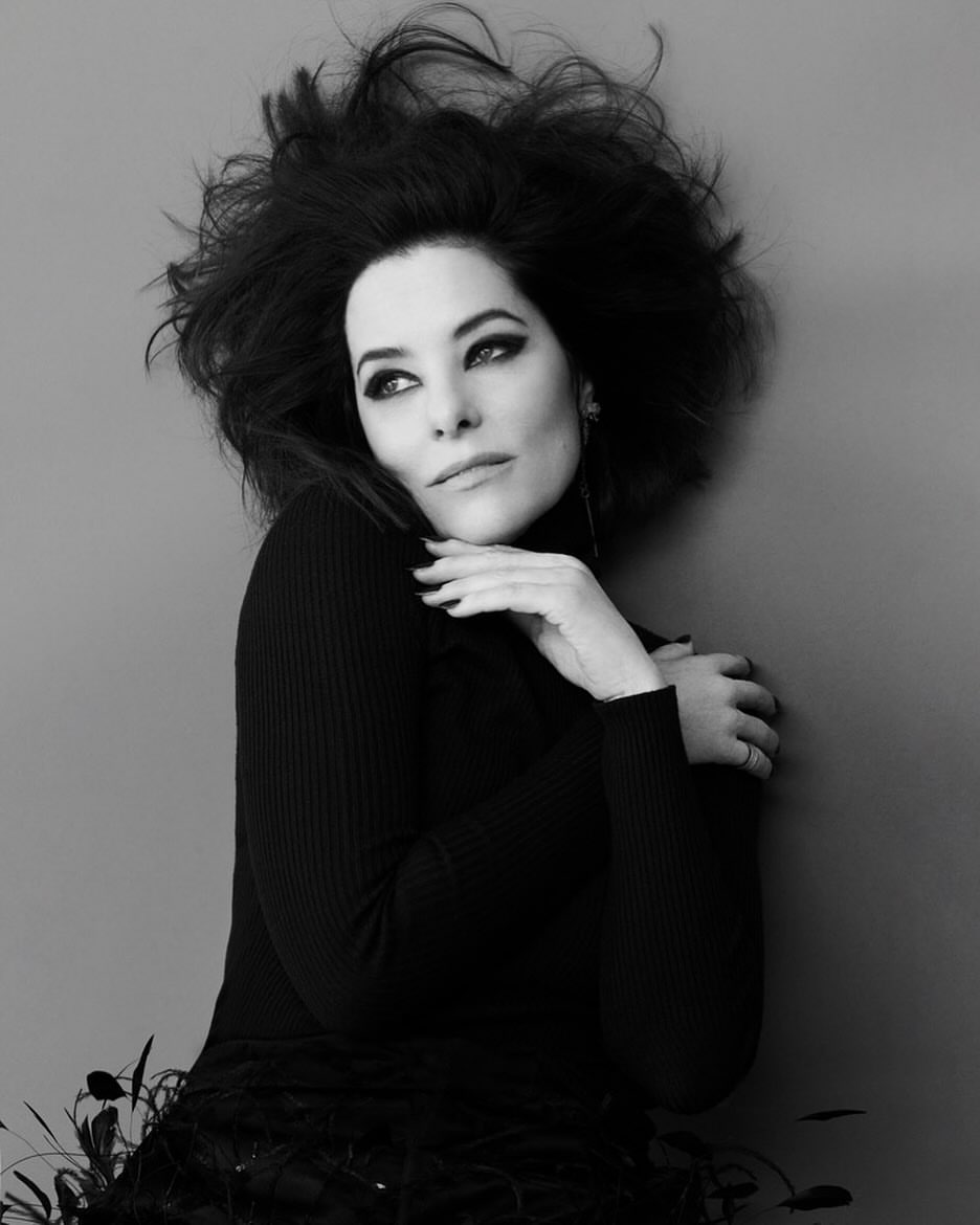 Black and white portrait of a brunette woman with dramatic hair and dark eye makeup in a black turtleneck sweater and feathered skirt.