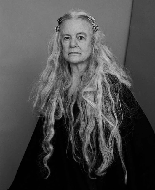 Monochrome portrait of an elderly woman with long, wavy, gray hair wearing a black garment, shot in a photo studio with a grey backdrop.
