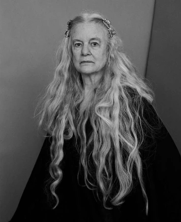 Monochrome portrait of an elderly woman with long, wavy, gray hair wearing a black garment, shot in a photo studio with a grey backdrop.