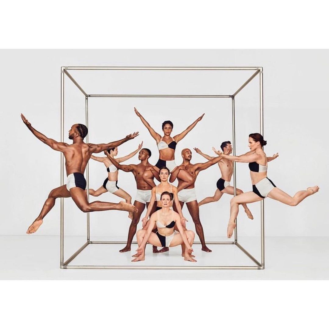 Eleven dancers wearing matching black and white outfits are leaping in a cube frame, set against a seamless white backdrop in a studio.
