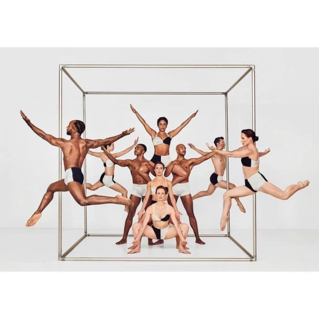 Eleven dancers wearing matching black and white outfits are leaping in a cube frame, set against a seamless white backdrop in a studio.