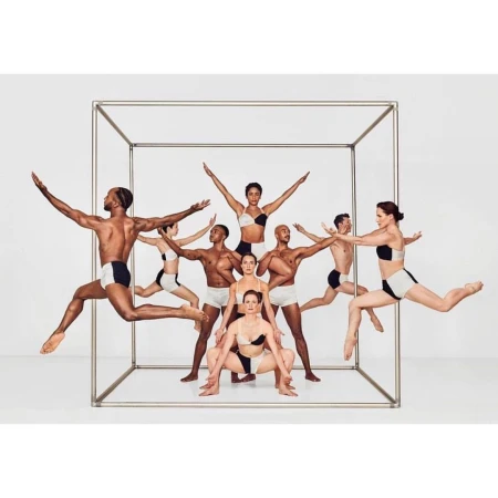 Eleven dancers wearing matching black and white outfits are leaping in a cube frame, set against a seamless white backdrop in a studio.