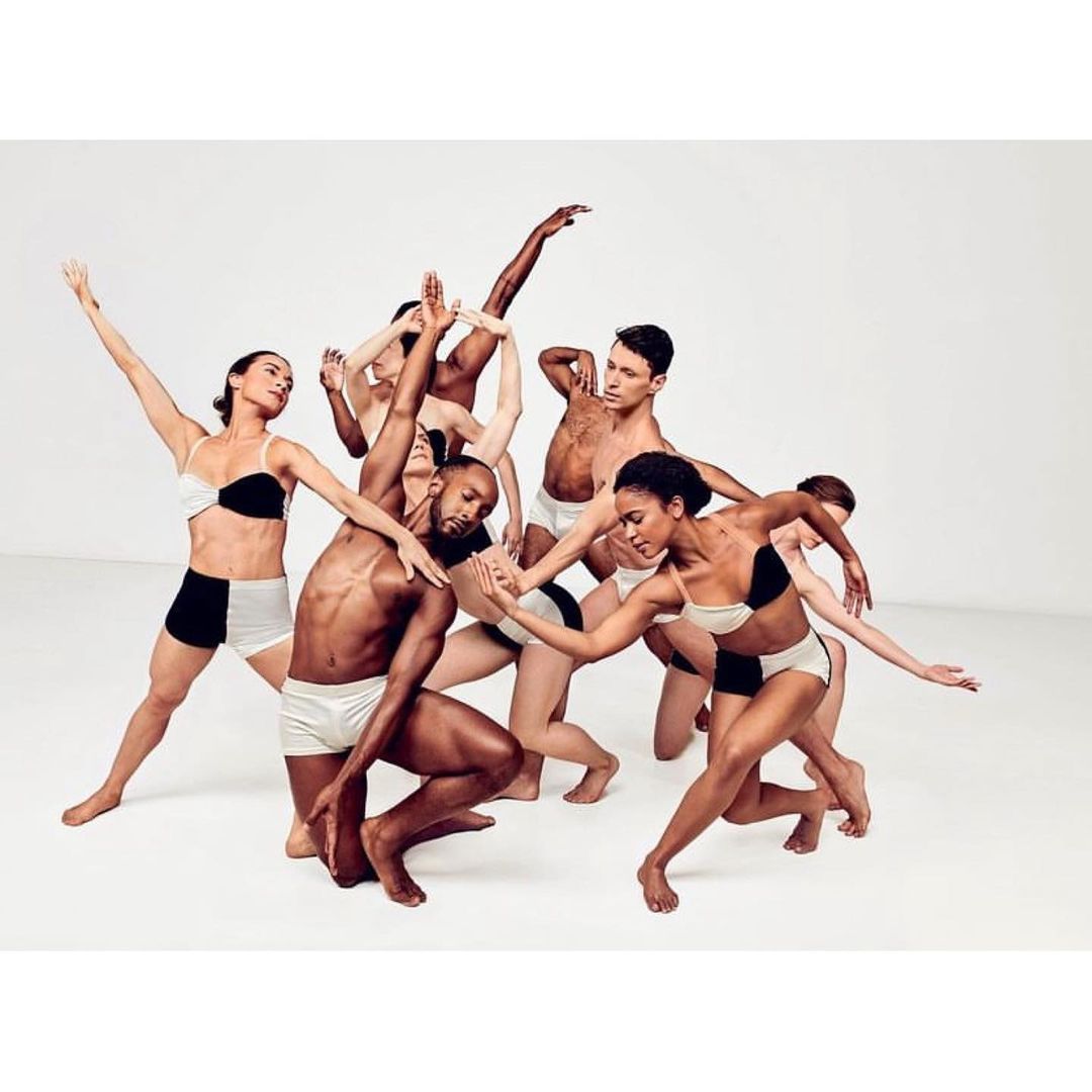 Group of nine diverse dancers in minimal clothing performing in a light studio setting with arms raised and legs bent, showcasing physical expression.