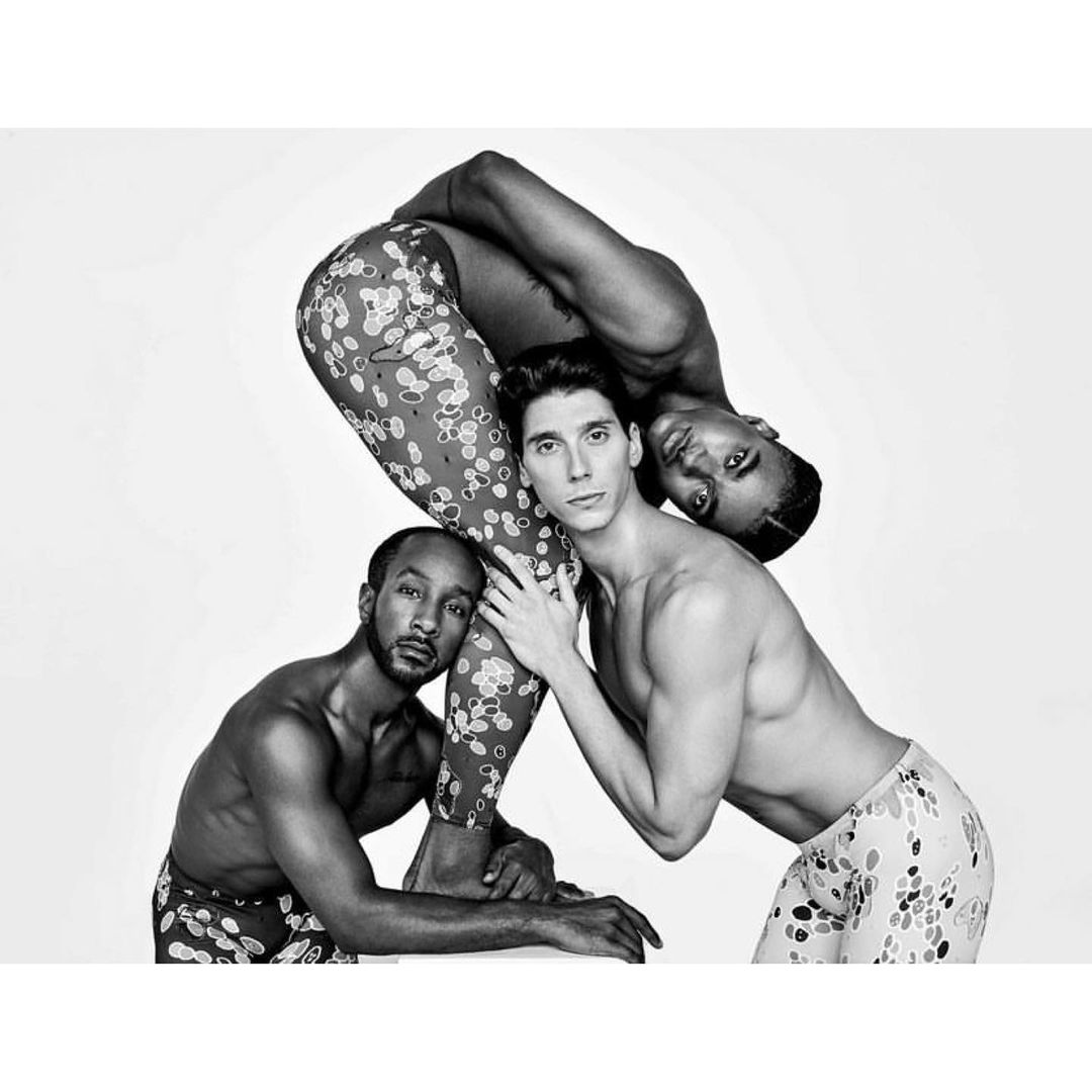 Three shirtless men in a striking pose against a white backdrop, featuring a mix of light skin and dark skin tones, and flower-print leggings.