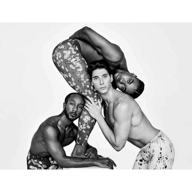 Three shirtless men in a striking pose against a white backdrop, featuring a mix of light skin and dark skin tones, and flower-print leggings.