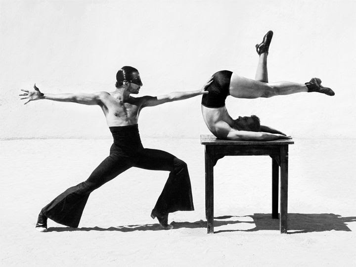Two athletic men performing a choreographed gymnastics routine on and around a small table in a bright, neutral-toned studio setting.