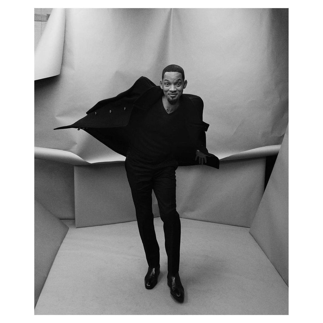 Monochrome full-body portrait of a smiling adult male in a black coat and pants posing in a boxy studio set with layered paper backdrop.