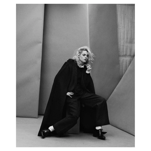 Monochrome studio portrait of a woman with blonde hair wearing a dark cape and wide-leg pants, seated against a backdrop.