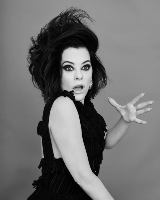 Black and white portrait of a woman with dark, voluminous hair and dramatic makeup, wearing a textured top against a plain background.