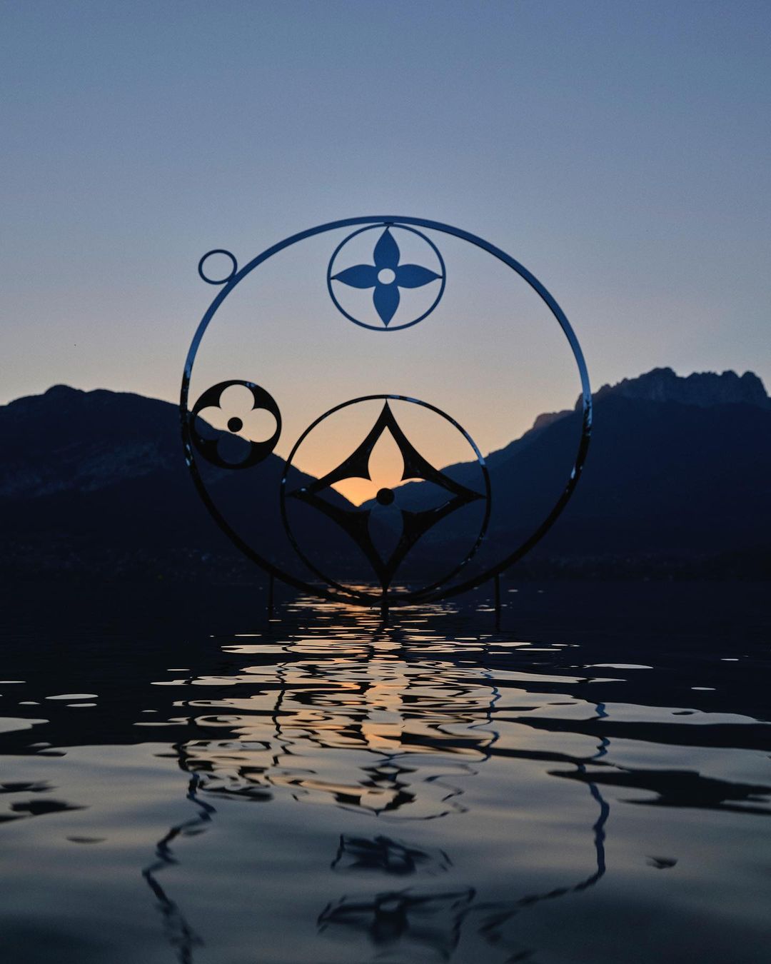 Abstract rendering of a logo reflected on water with a mountain range and twilight sky in the background, creating a serene atmosphere.