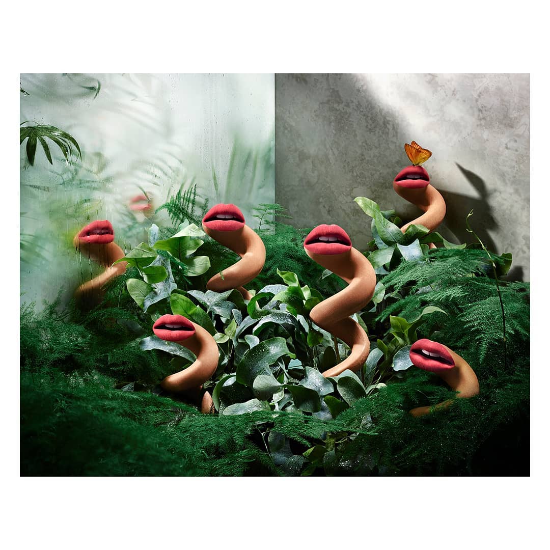 Surreal image of human lips on fleshy stalks in a verdant botanical setting with a butterfly perched on one of the lips.