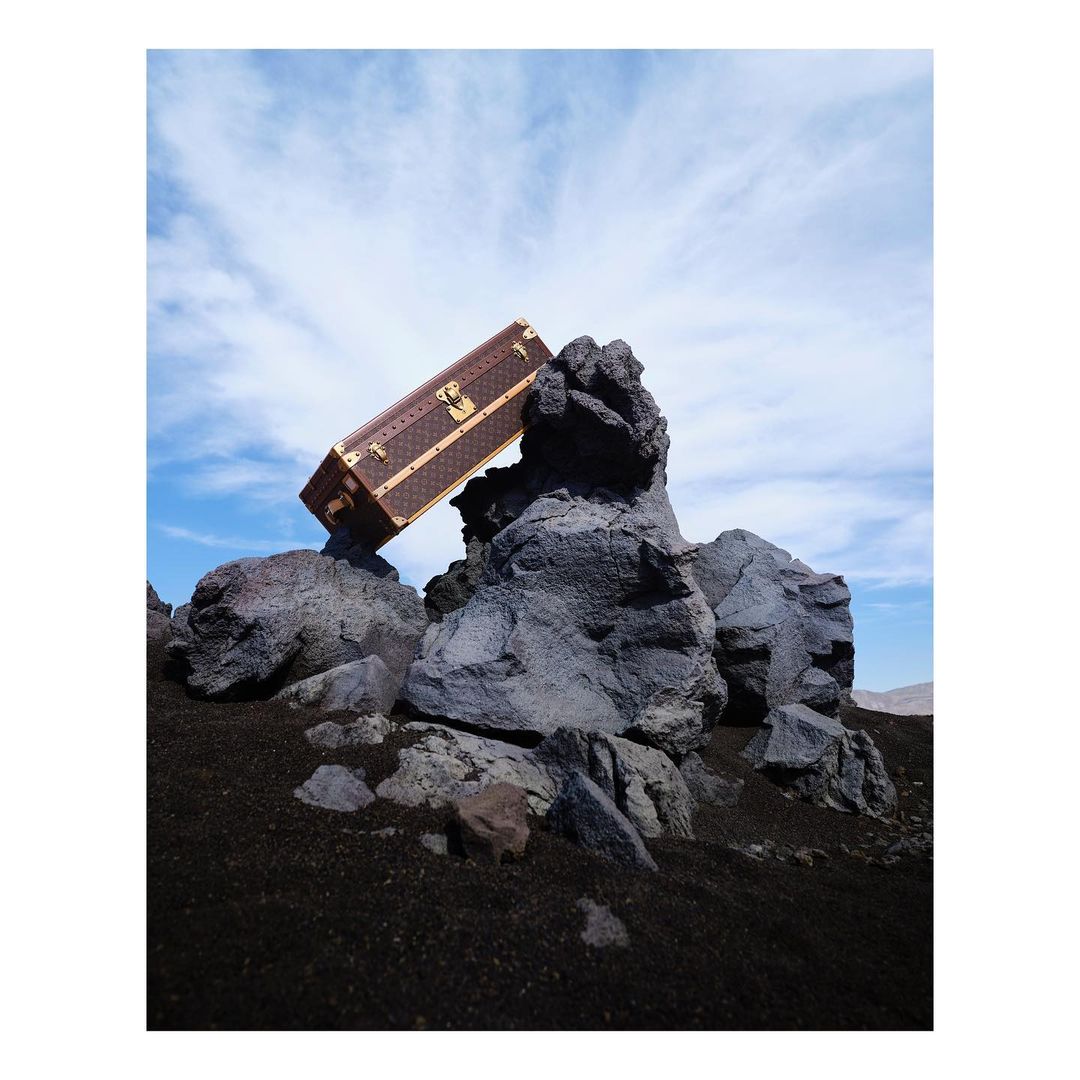 Brown Louis Vuitton trunk rests on a rocky outcrop against a sky of blue and white clouds. Luggage is ready for travel.