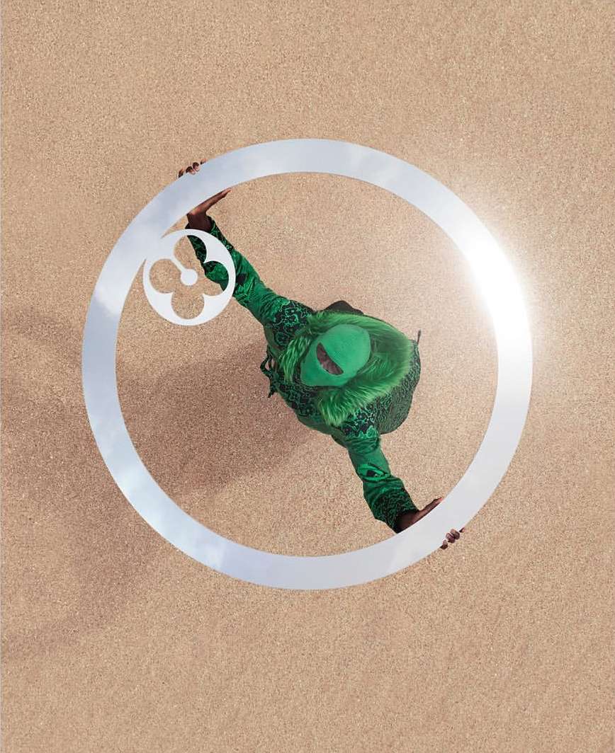 Overhead shot of a person in green patterned clothing standing inside a reflective ring on a sandy surface with bright sunlight.
