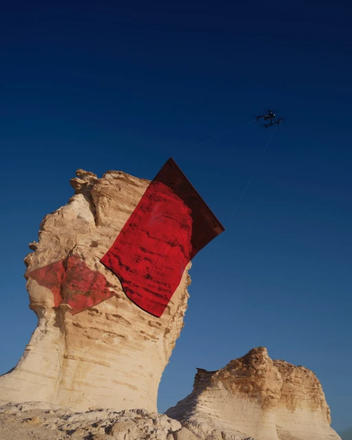 Drone flies in clear blue sky over tan rock formations in the desert with a red flag obscuring the rock face in the daylight.