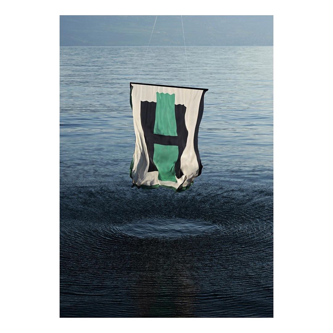 Abstract artwork featuring a cloth banner with a green 'H' design suspended over a serene body of water with mountains in the distance.