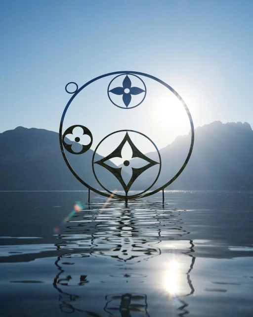 Metal sculpture with floral patterns standing in rippled water, set against a backdrop of mountains and a bright, clear sky.