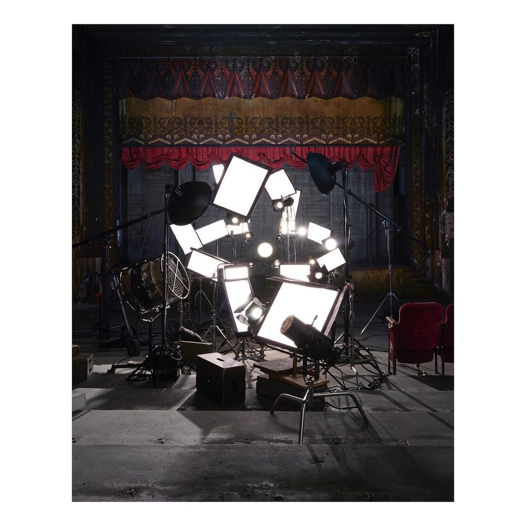 Indoor shot of theater stage with visible red stage curtains, numerous stage lighting stands, vintage metal fan, chair, and stage floor.