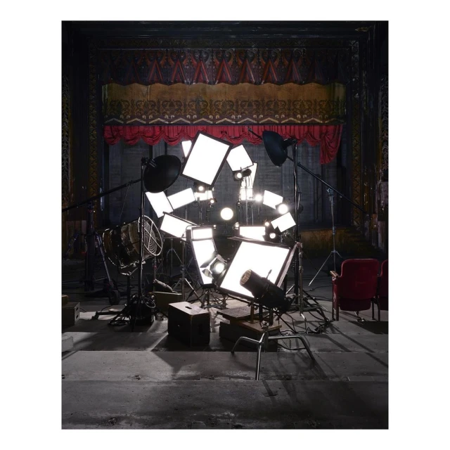 Indoor shot of theater stage with visible red stage curtains, numerous stage lighting stands, vintage metal fan, chair, and stage floor.