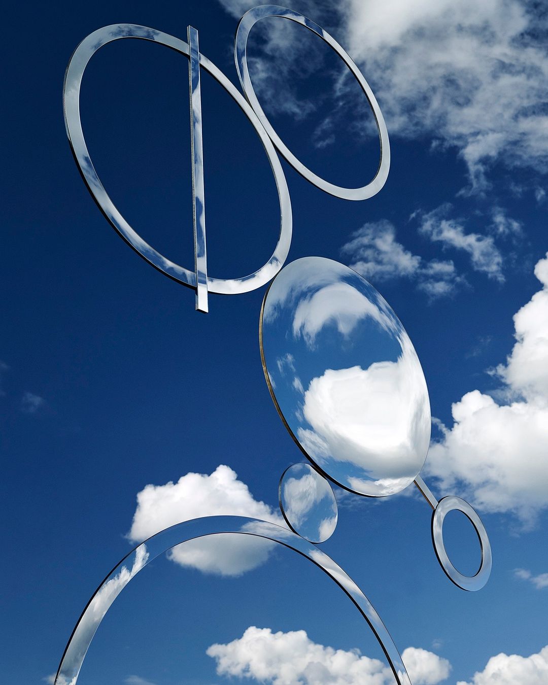 Abstract metal sculpture of reflective rings and lines against a bright blue sky with white clouds creating a contemporary art display.