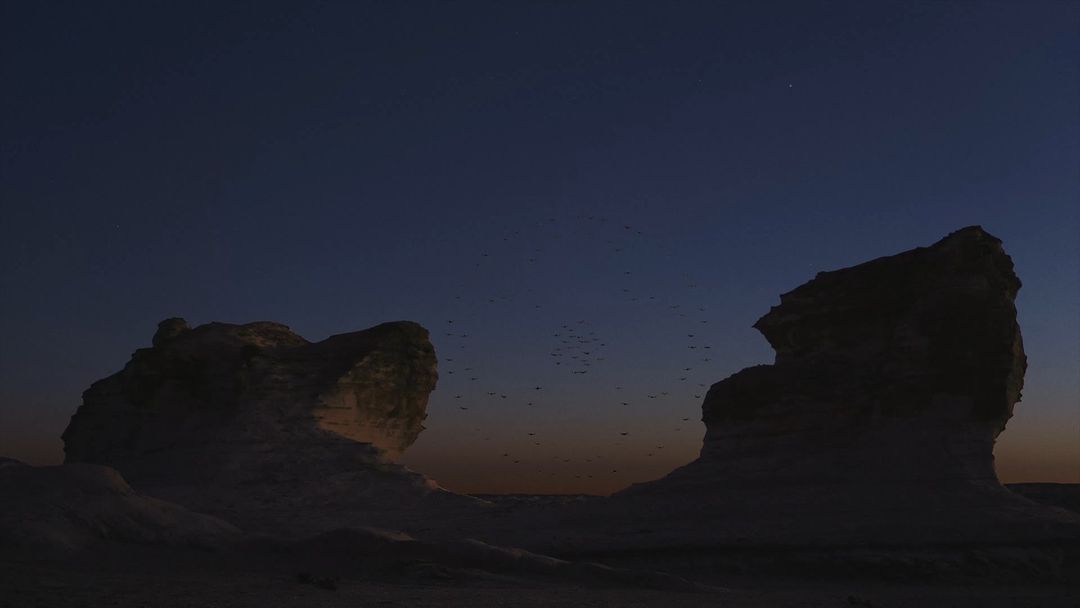Desert landscape at dusk featuring two large rock formations silhouetted against a gradient sky with a flock of birds flying between them.