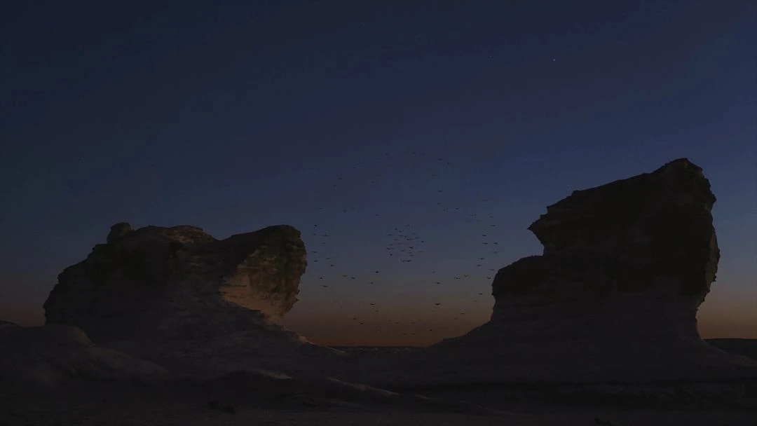 Desert landscape at dusk featuring two large rock formations silhouetted against a gradient sky with a flock of birds flying between them.