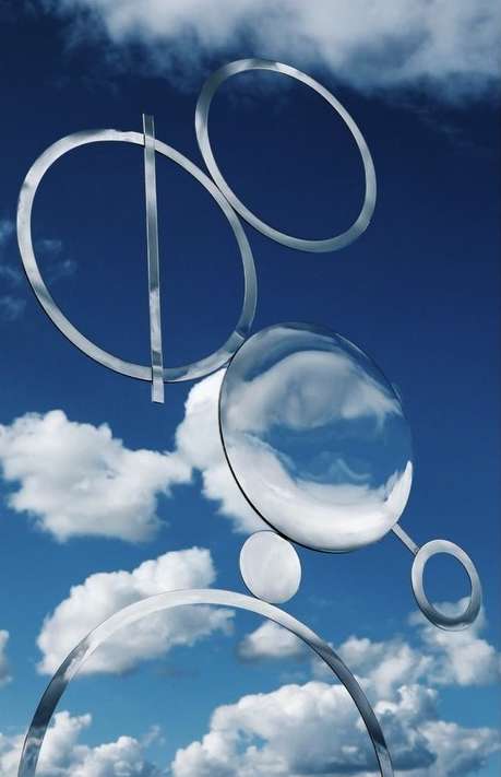 Abstract chrome metal sculpture featuring circles and spheres set against a vibrant blue sky with fluffy white clouds in the background.