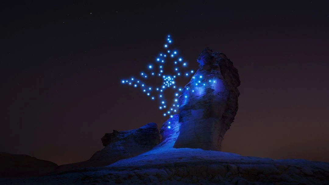 Illuminated rock formation at night under a dark sky with a display of arranged drones creating a glowing light pattern.
