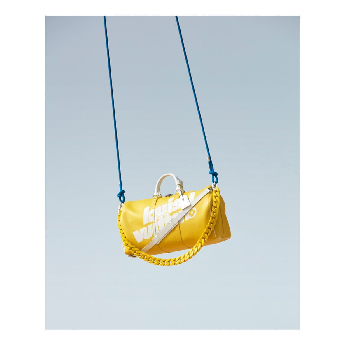 Yellow Louis Vuitton duffle bag with white logo and chain strap, suspended by blue cords against a bright blue sky.
