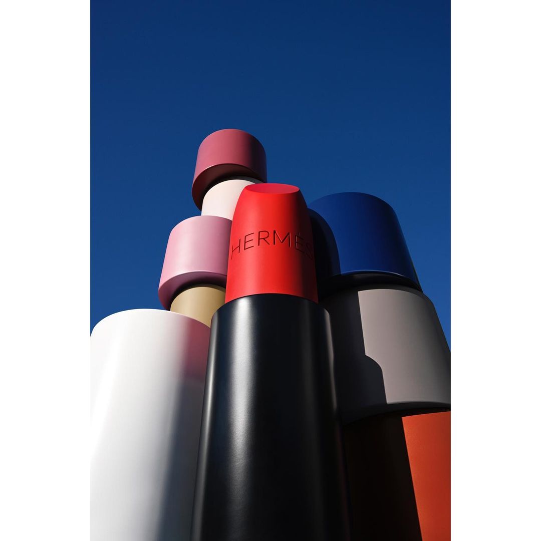 Group of oversized lipstick tubes in varying colors against bright blue sky, featuring the Hermès logo on one tube.