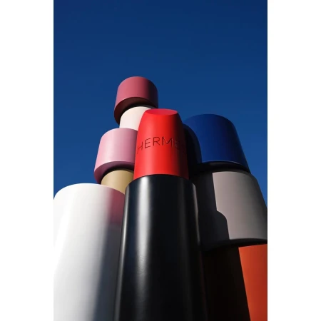 Group of oversized lipstick tubes in varying colors against bright blue sky, featuring the Hermès logo on one tube.
