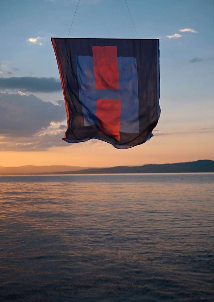 Abstract fabric banner in red and blue hangs against a vivid sky and reflective ocean at sunset, creating a surreal seascape.
