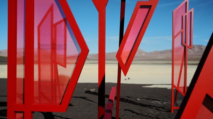 Abstract red geometric sculpture on a desert landscape with a clear blue sky and distant mountains. Daytime setting with bright light.