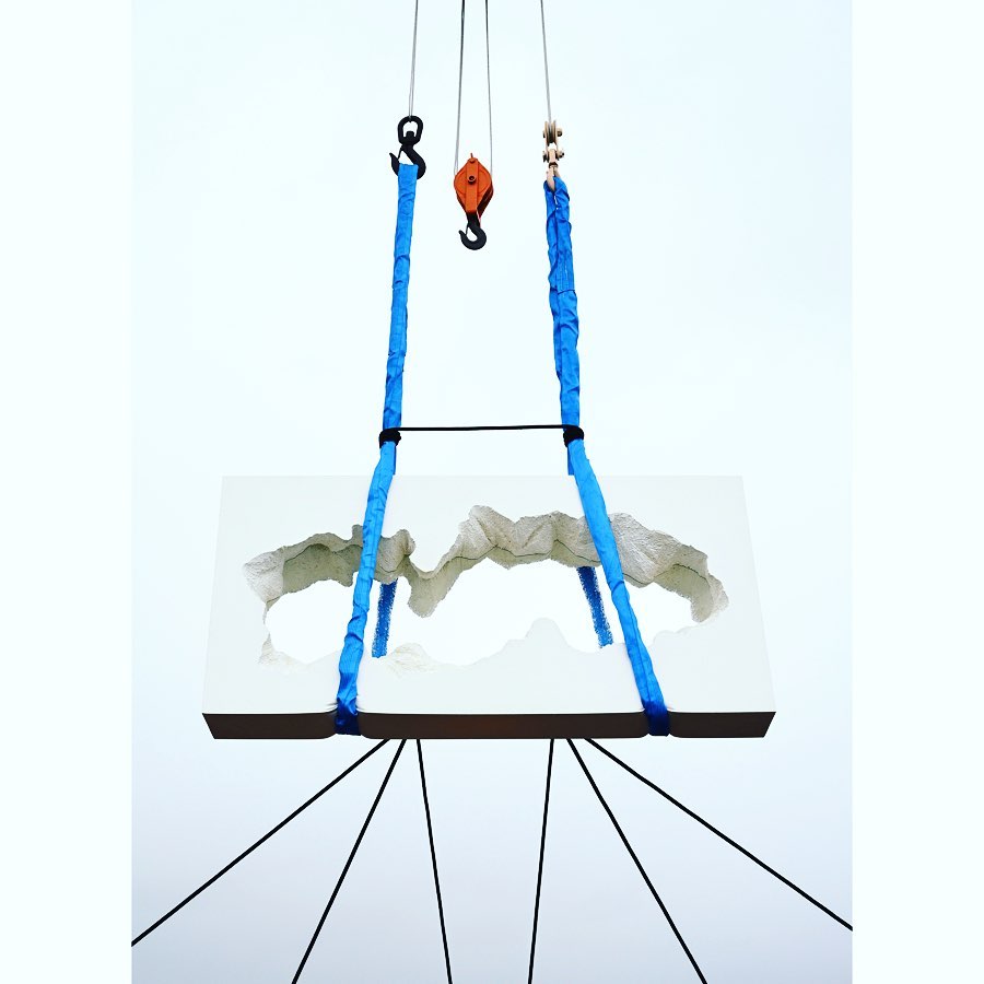 White rectangular object with interior landscape design hanging from blue straps and black cables against a light neutral background.