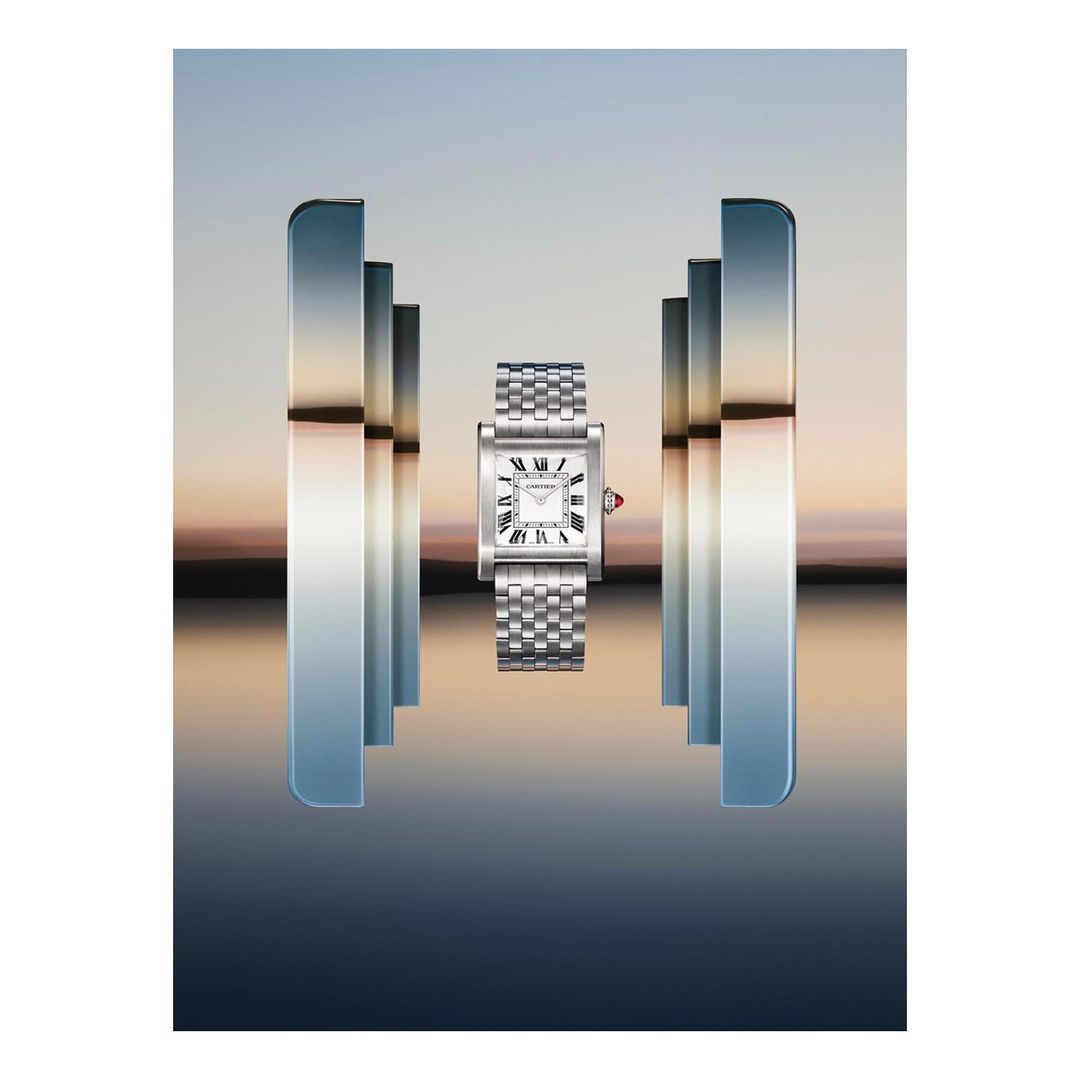 Silver Cartier Tank watch on a metal bracelet, presented with vertical bars of reflective chrome, set against a blurred sunset sky.