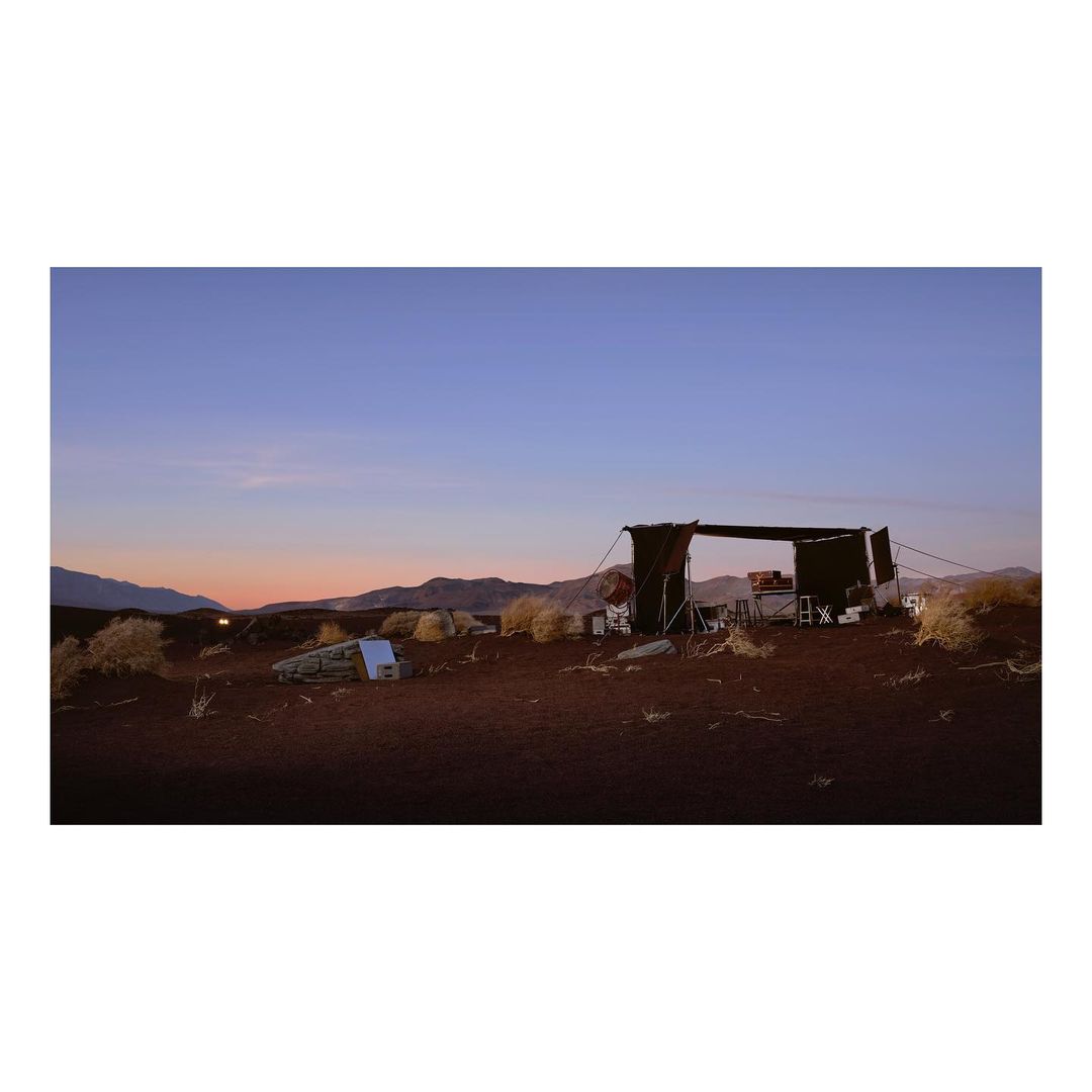 Open-air desert shelter at dusk. Shelter with chairs and tables surrounded by desert landscape, bushes, and mountains in the background.