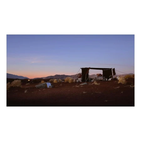 Open-air desert shelter at dusk. Shelter with chairs and tables surrounded by desert landscape, bushes, and mountains in the background.