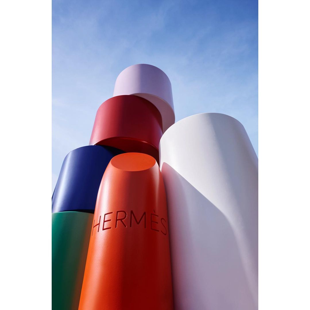 Stack of large lipstick tubes in vibrant colors with the word HERMES on one, against a bright blue sky backdrop.