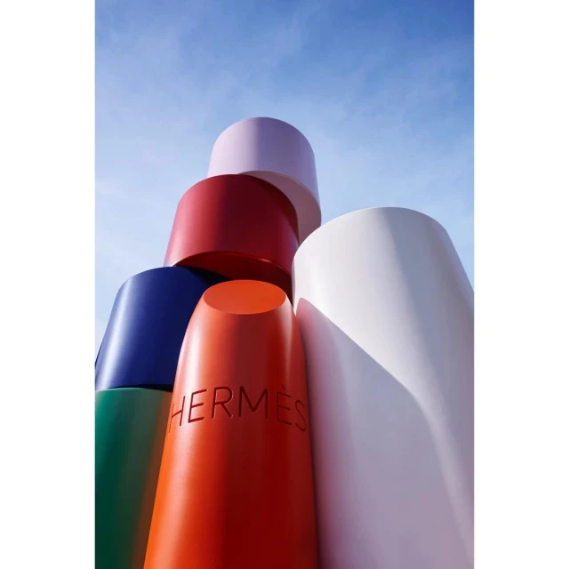 Stack of large lipstick tubes in vibrant colors with the word HERMES on one, against a bright blue sky backdrop.