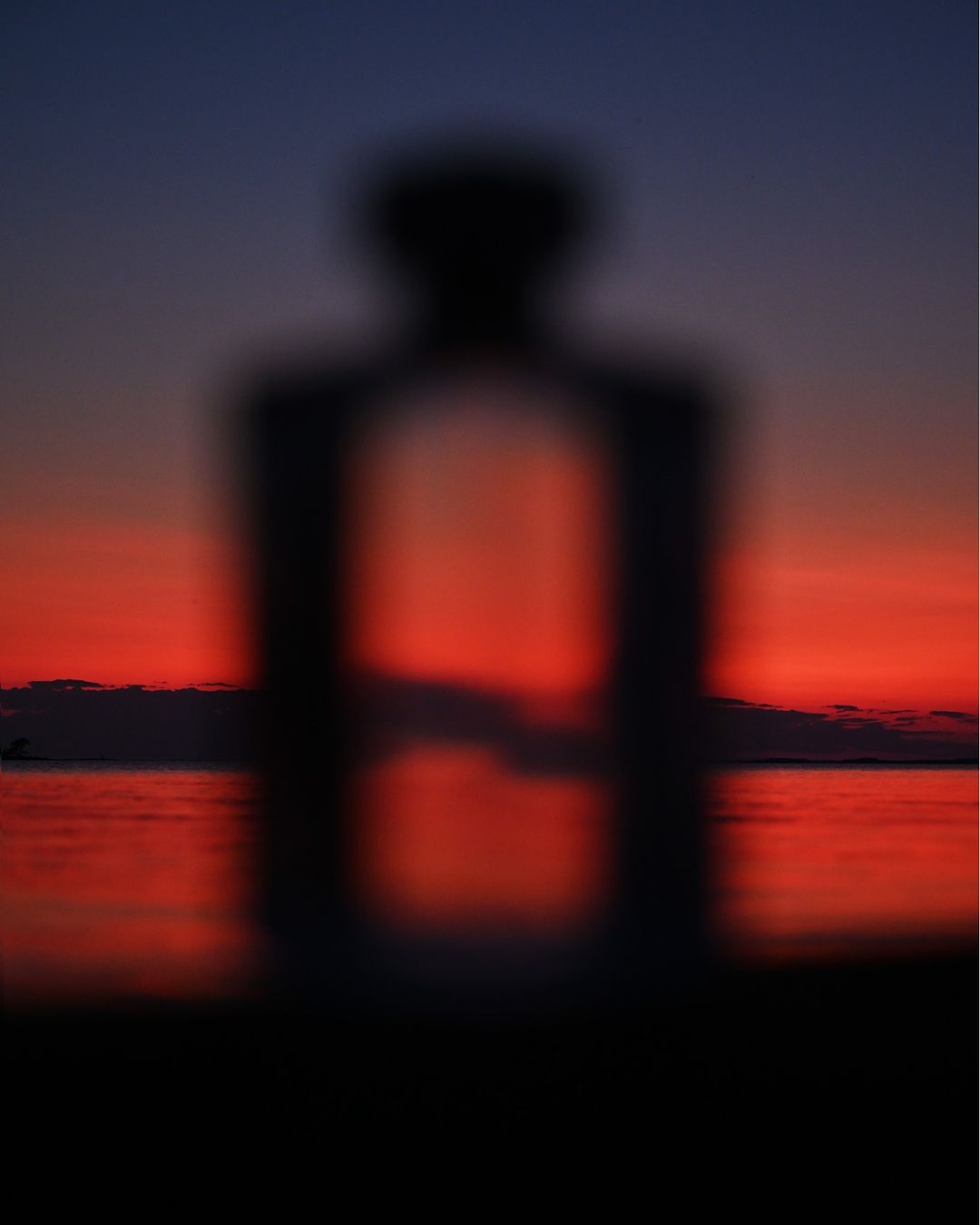 Blurred figure silhouette against a sunset over water. The sky transitions from orange to dark blue, with the sun reflecting on the sea.