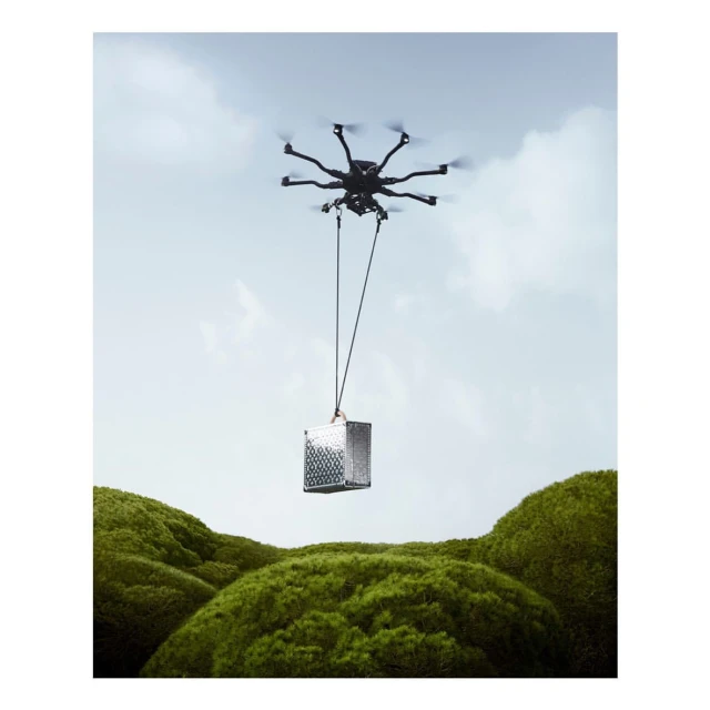 Delivery drone with a silver cube package suspended by thin cables, against a pale blue sky and green, soft-shaped hills.