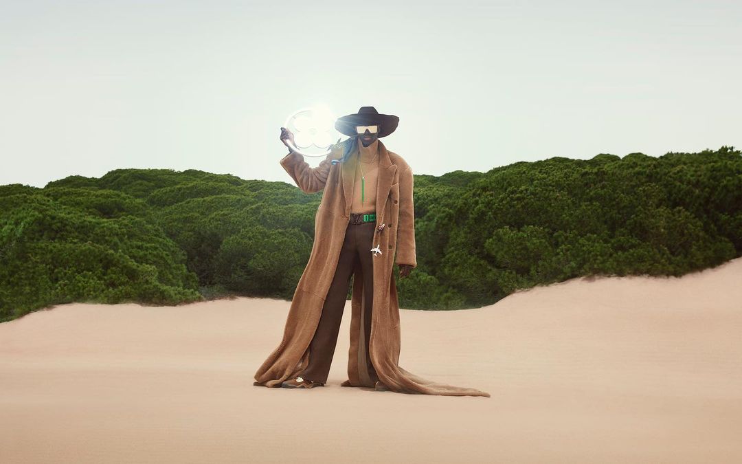 Fashion shoot featuring an adult male model wearing a long brown coat and sunglasses, holding a circular light in a desert landscape.