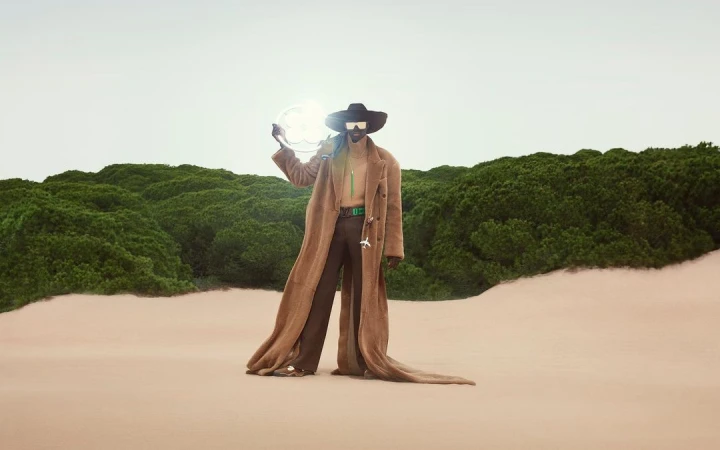 Fashion shoot featuring an adult male model wearing a long brown coat and sunglasses, holding a circular light in a desert landscape.