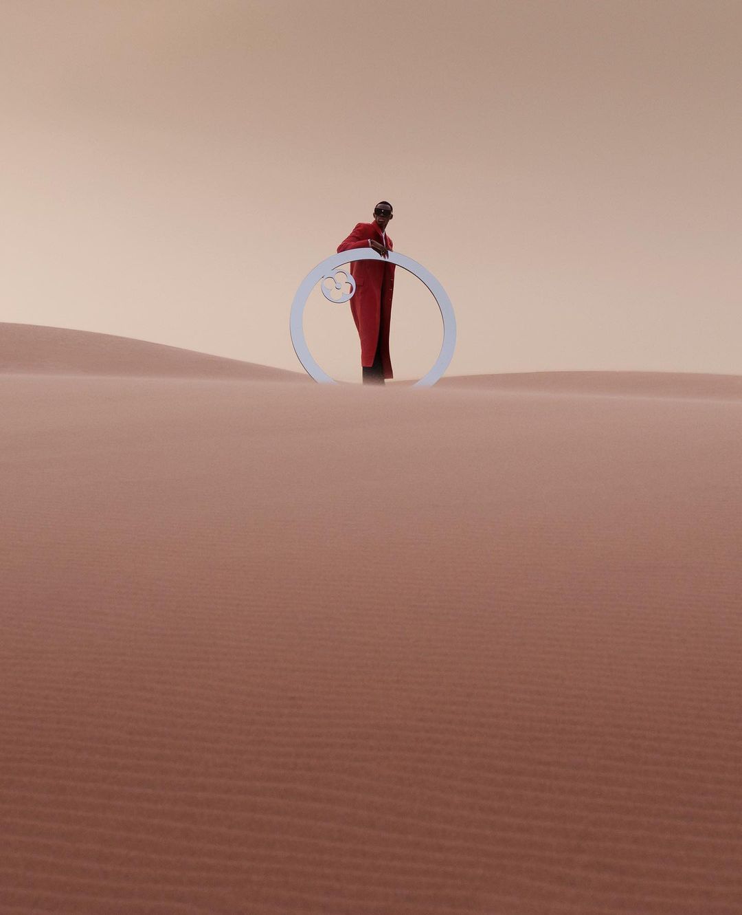 A single person in a red robe stands with a circular white prop in a minimalist desert landscape with orange-tinted sky and sand dunes.