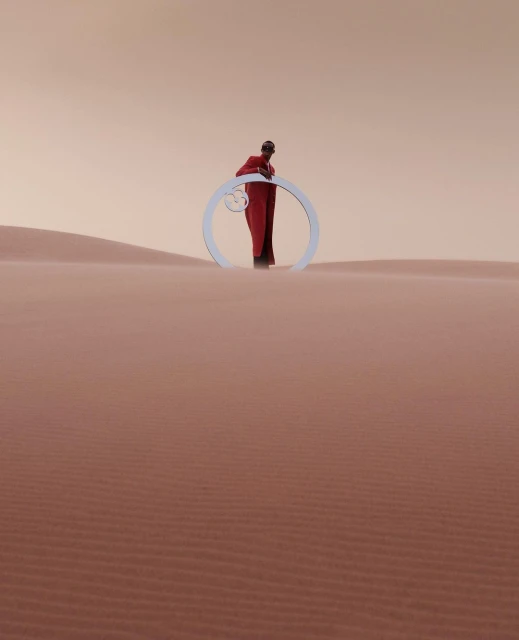 A single person in a red robe stands with a circular white prop in a minimalist desert landscape with orange-tinted sky and sand dunes.