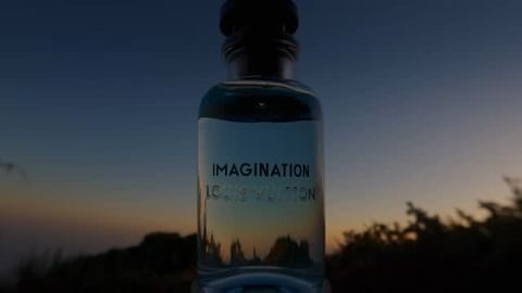 Close-up of a clear glass Louis Vuitton fragrance bottle in front of a twilight sky with blurry trees on the horizon.