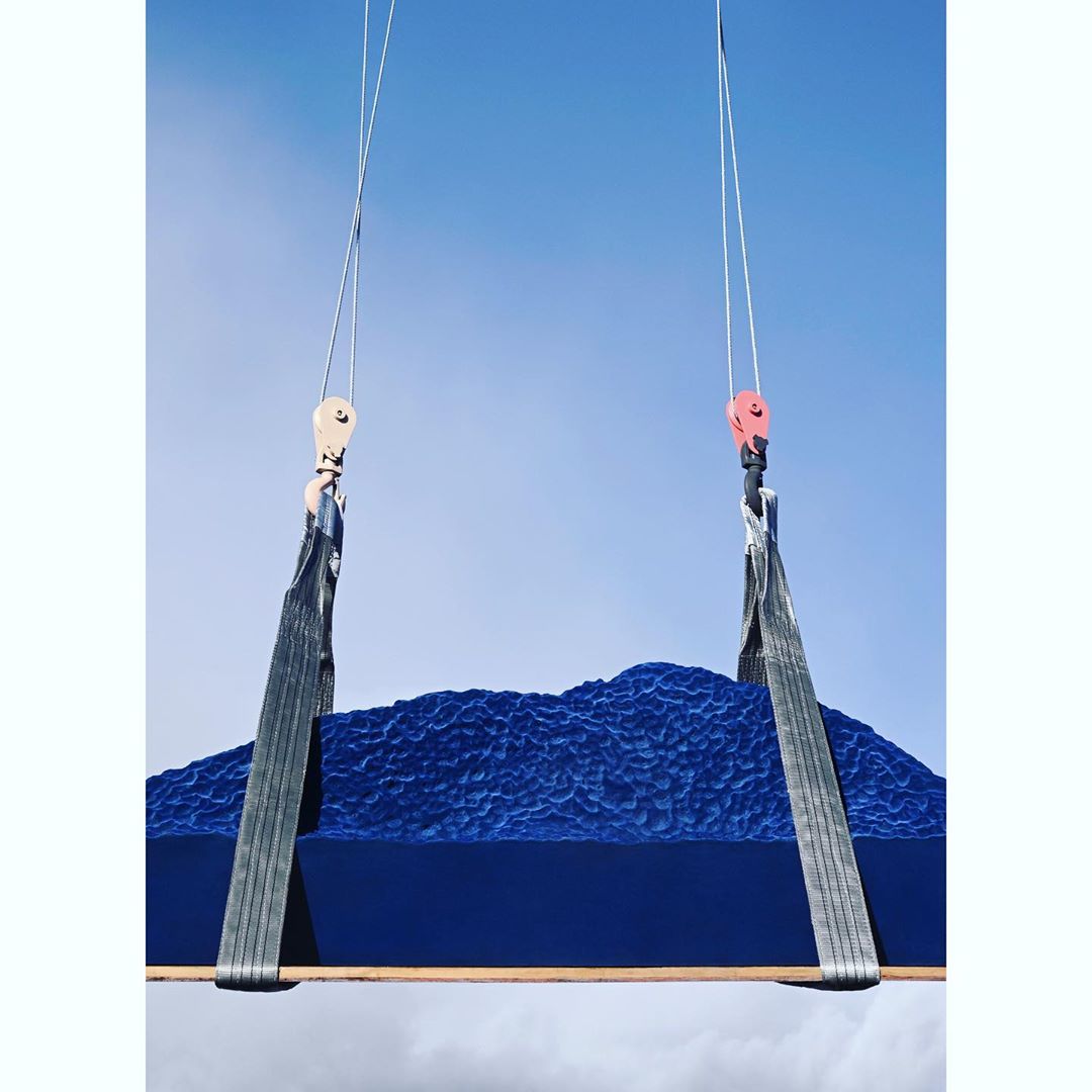Blue abstract art piece with wave-like texture suspended by industrial straps against a clear blue sky. Installation art.